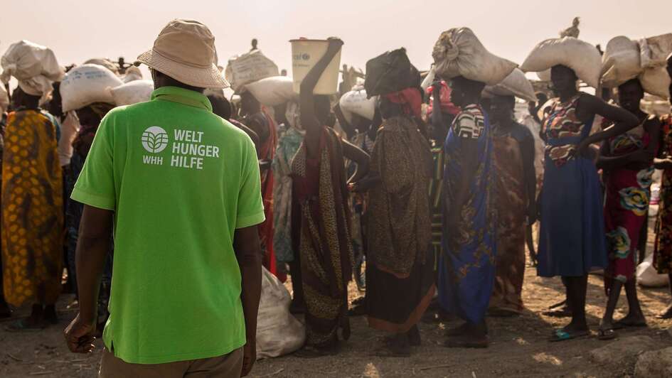 Mitarbeiter der Welthungerhilfe in einem grünen T-Shirt mit dem neu gestalteten Welthungerhilfe-Logo. Im Hintergrund sind Menschen zu sehen, die Dinge auf ihren Köpfen tragen.