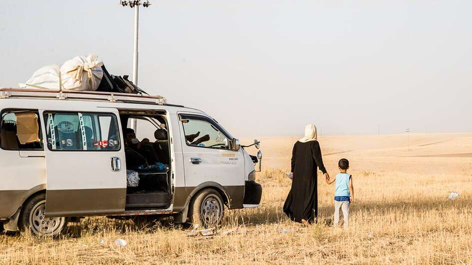 Frau und Kind blicken in die Ferne, daneben ist ein VW-Omnibus ähnliches Auto.