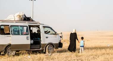 Frau und Kind blicken in die Ferne, daneben ist ein VW-Omnibus ähnliches Auto.