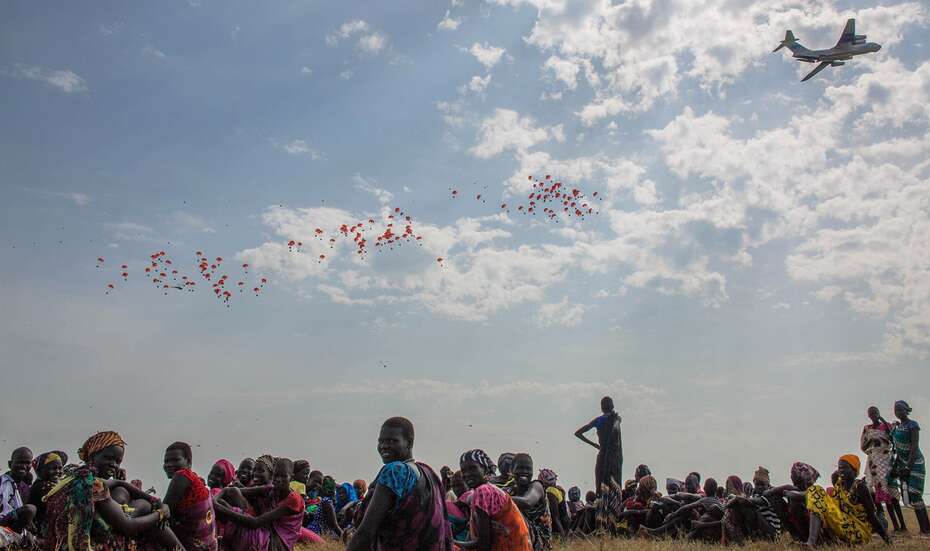 Auf dem Boden sitzen Menschen auf einem Feld, in der Ferne sieht man ein Flugzeug, das Nahrungsmittel aus der Luft ablädt.