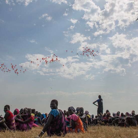 Auf dem Boden sitzen Menschen auf einem Feld, in der Ferne sieht man ein Flugzeug, das Nahrungsmittel aus der Luft ablädt.
