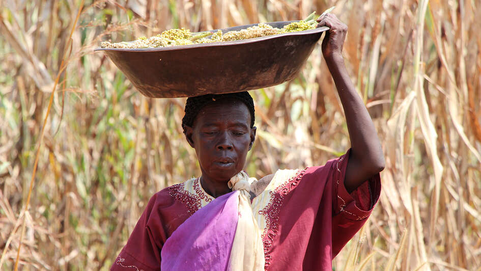 Eine Frau im Südsudan steht - einen Korb auf ihrem Kopf tragend - vor einem Feld.